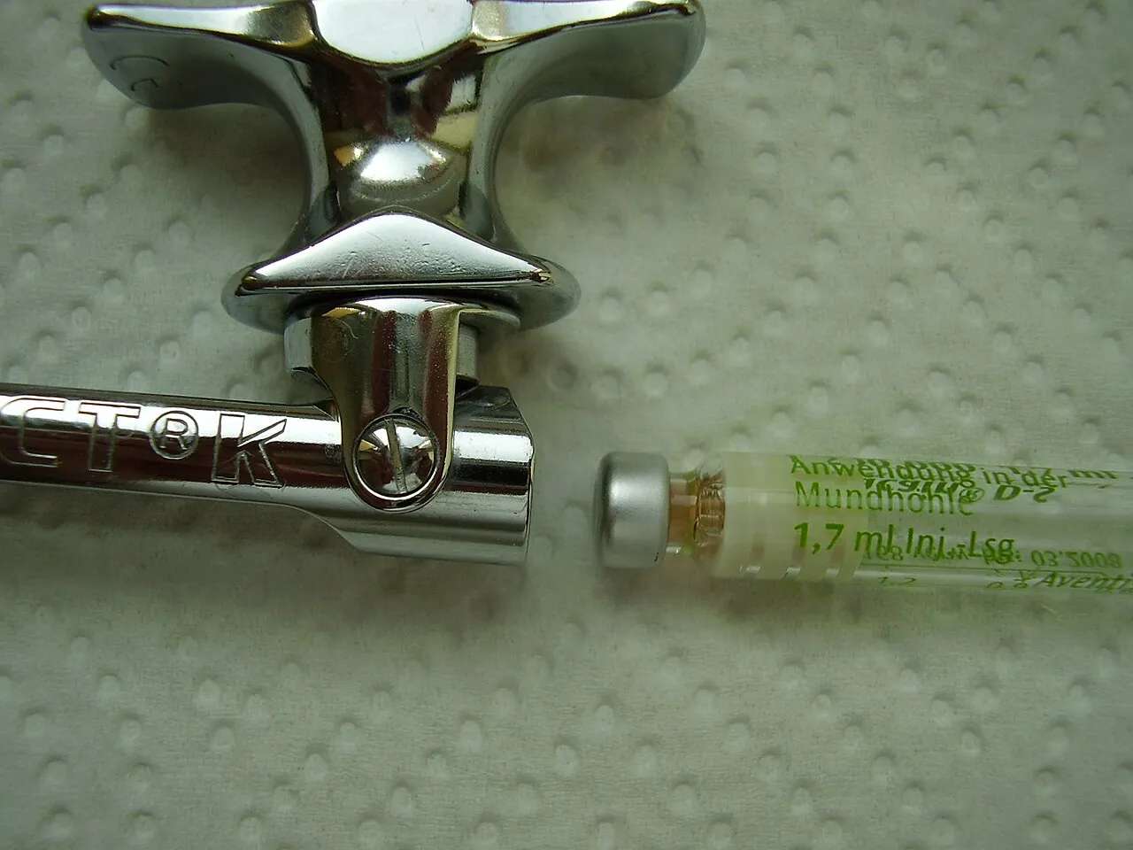 A 1.7 mL anesthetic cartridge lying beside the barrel and thumb ring of a dental cartridge syringe