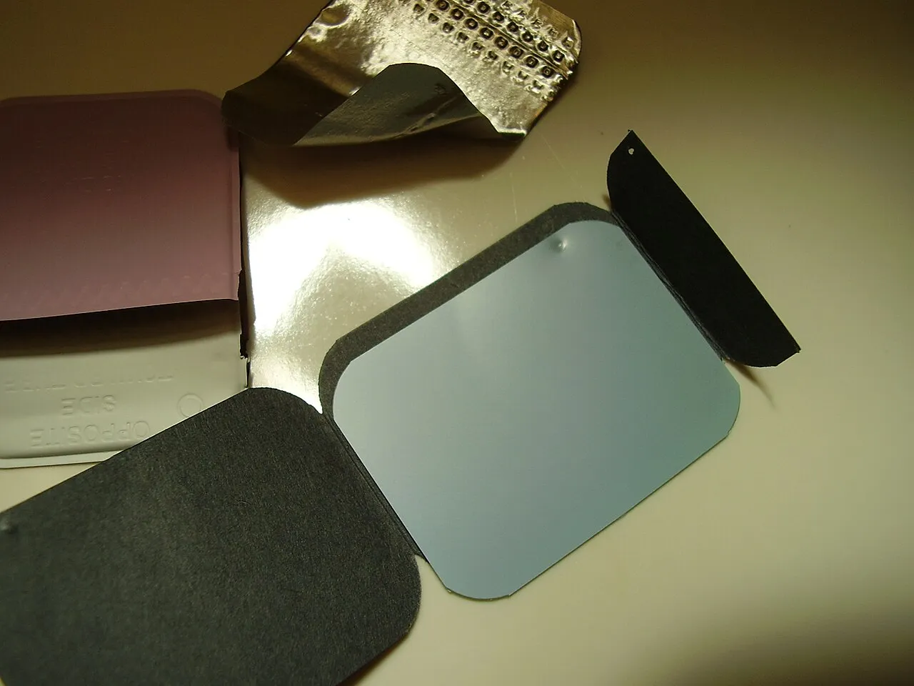An opened intraoral film packet laid out in its layers: the outer wrap marked opposite side toward tube, the black protective paper, the film itself and the embossed lead foil