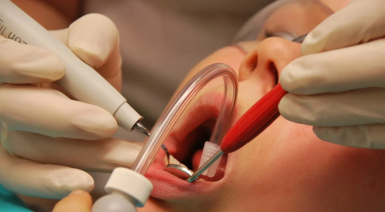 A clinician uses a power scaler on a patient wearing protective eyewear while an assistant holds a suction tip and a mirror to control water and aerosol